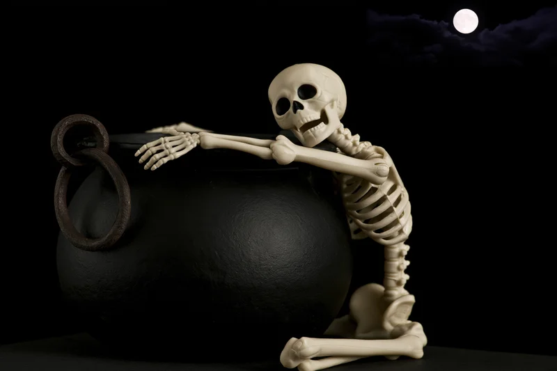 Small plastic skeleton figurine draped over black cauldron against night sky with full moon
