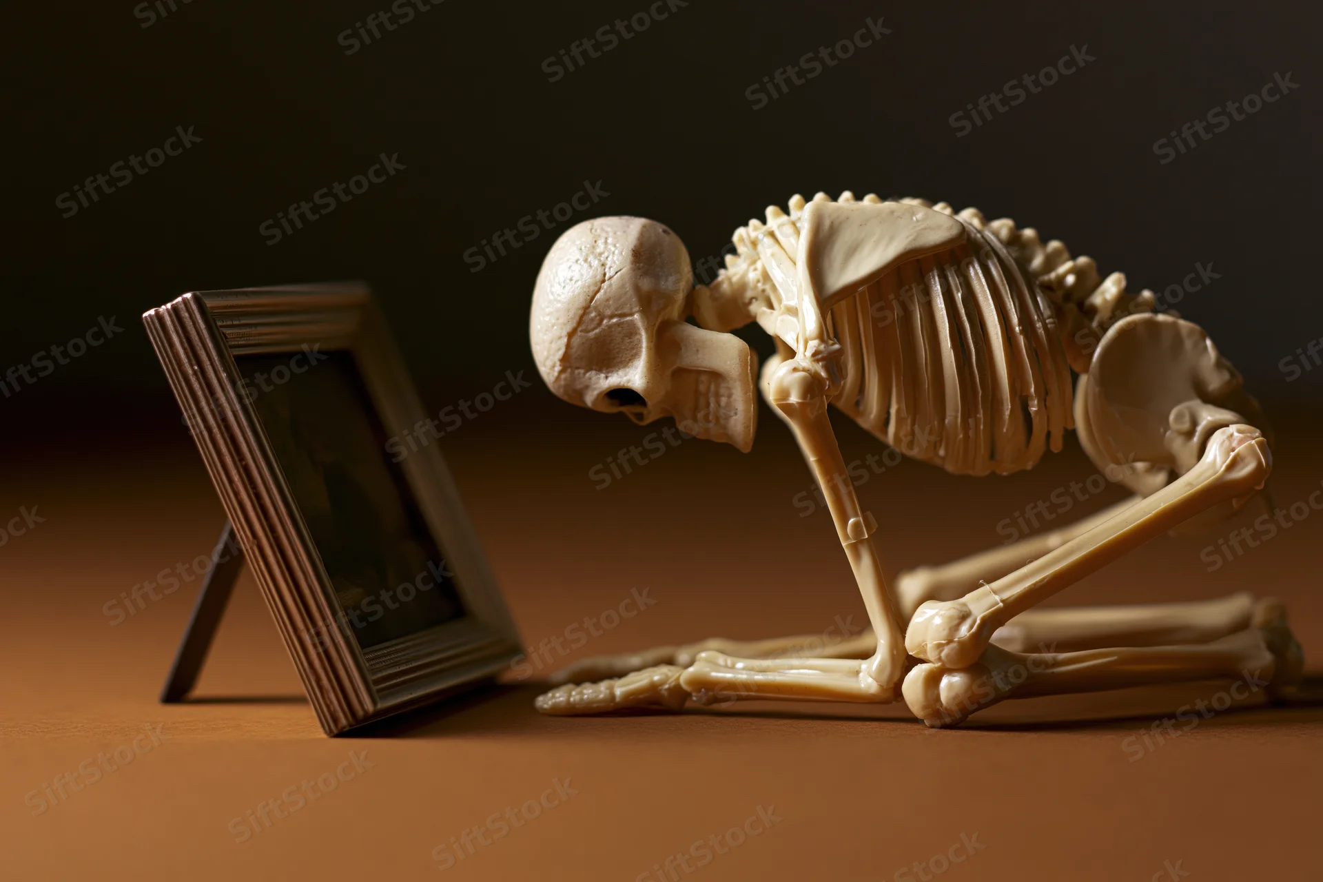 Small plastic human skeleton figurine leaning forward on a brown surface beside a framed picture