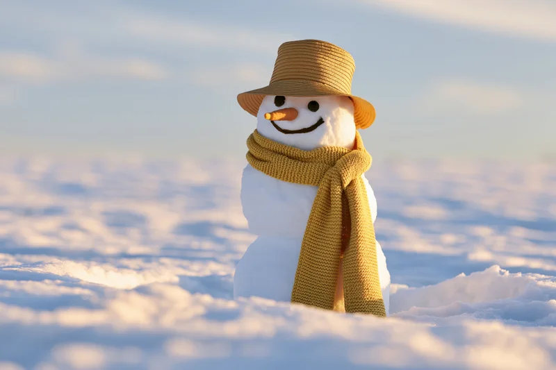 Small snowman with carrot nose and yellow hat and scarf standing in snowfield under blue sky