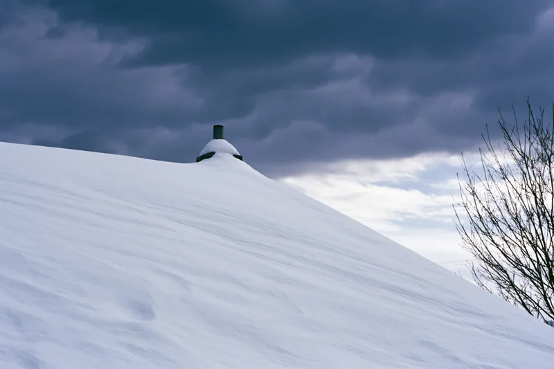 Snow-covered chimney on a sloped hill under stormy sky with bare tree branches visible