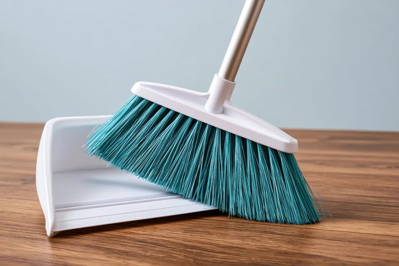 Teal-bristled broom resting against white plastic dustpan on wooden floor