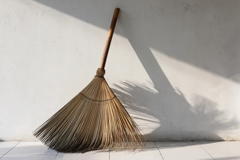 Traditional coconut-spine broom leaning against white wall in sunlit interior