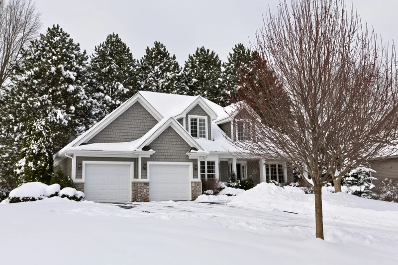 Snow-covered suburban house with gray cedar shake siding and two-car garage in winter landscape