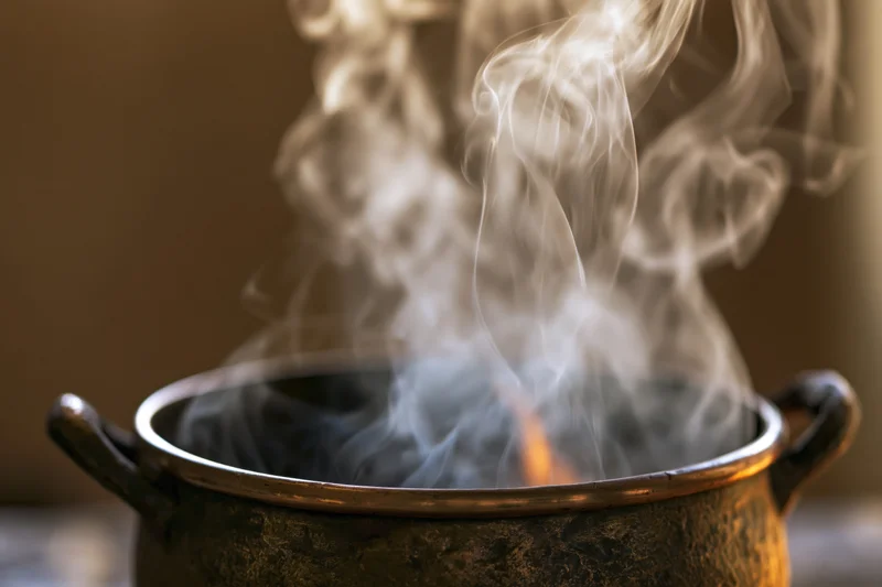 Steaming cauldron with fire inside, emitting smoke, on a dark surface