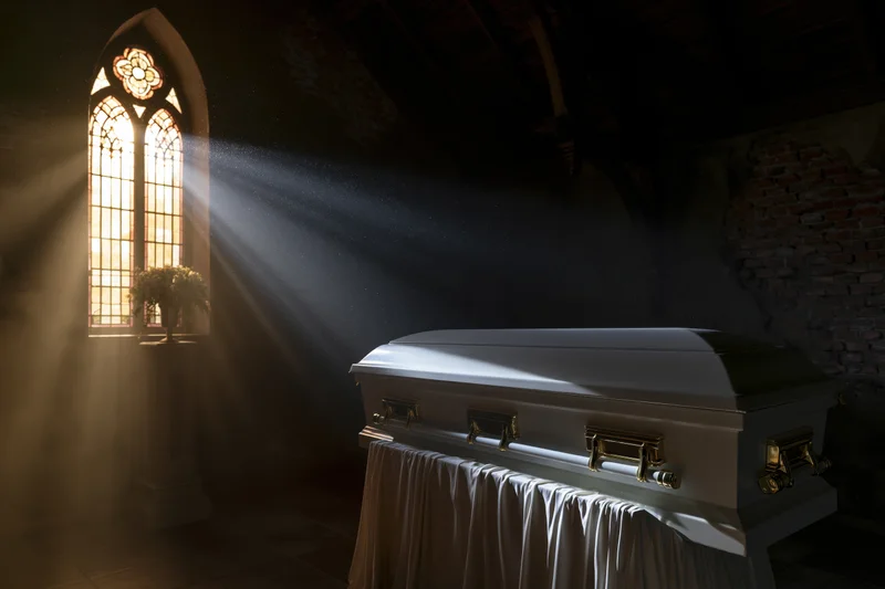 White casket with gold hardware on draped stand in church interior lit by stained glass window rays