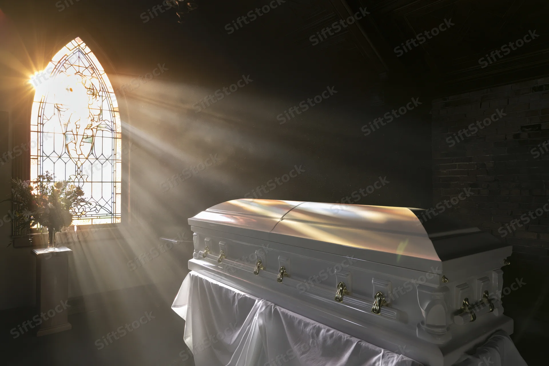 White casket with gold hardware on draped stand in church lit by stained glass window sunlight