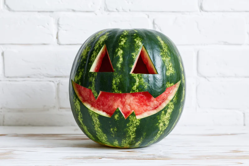 Carved watermelon with jack-o-lantern face on white wooden table against brick wall