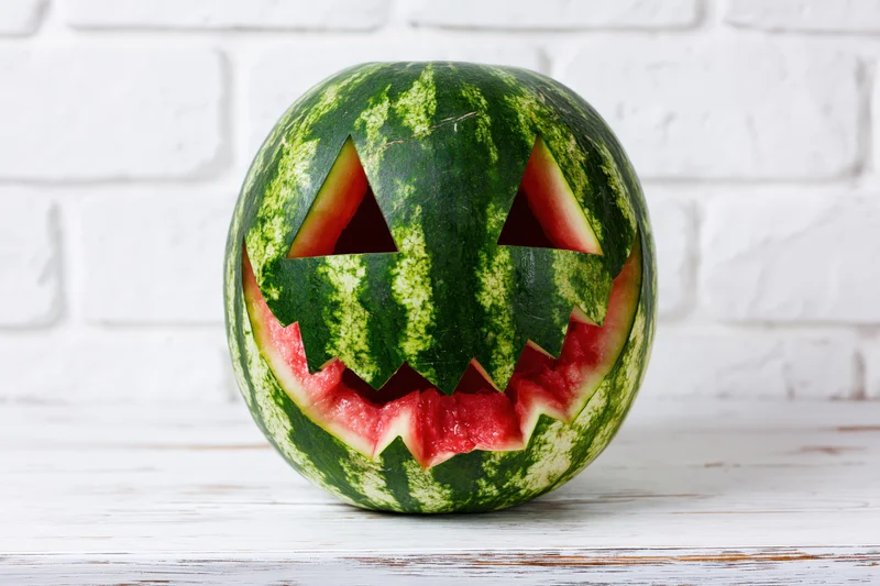 Carved watermelon with jack-o-lantern face on white wooden table against brick wall