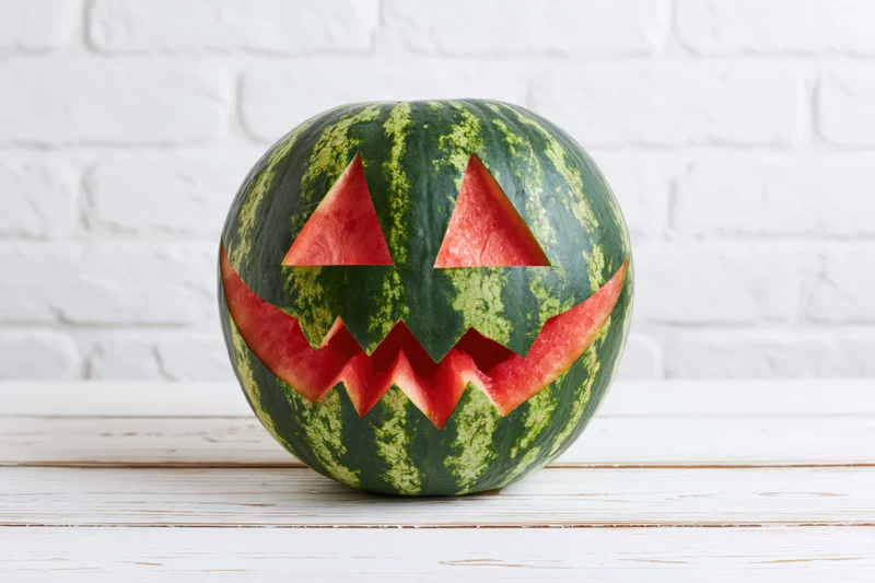 Carved watermelon with jack-o-lantern face on white wooden table against brick wall