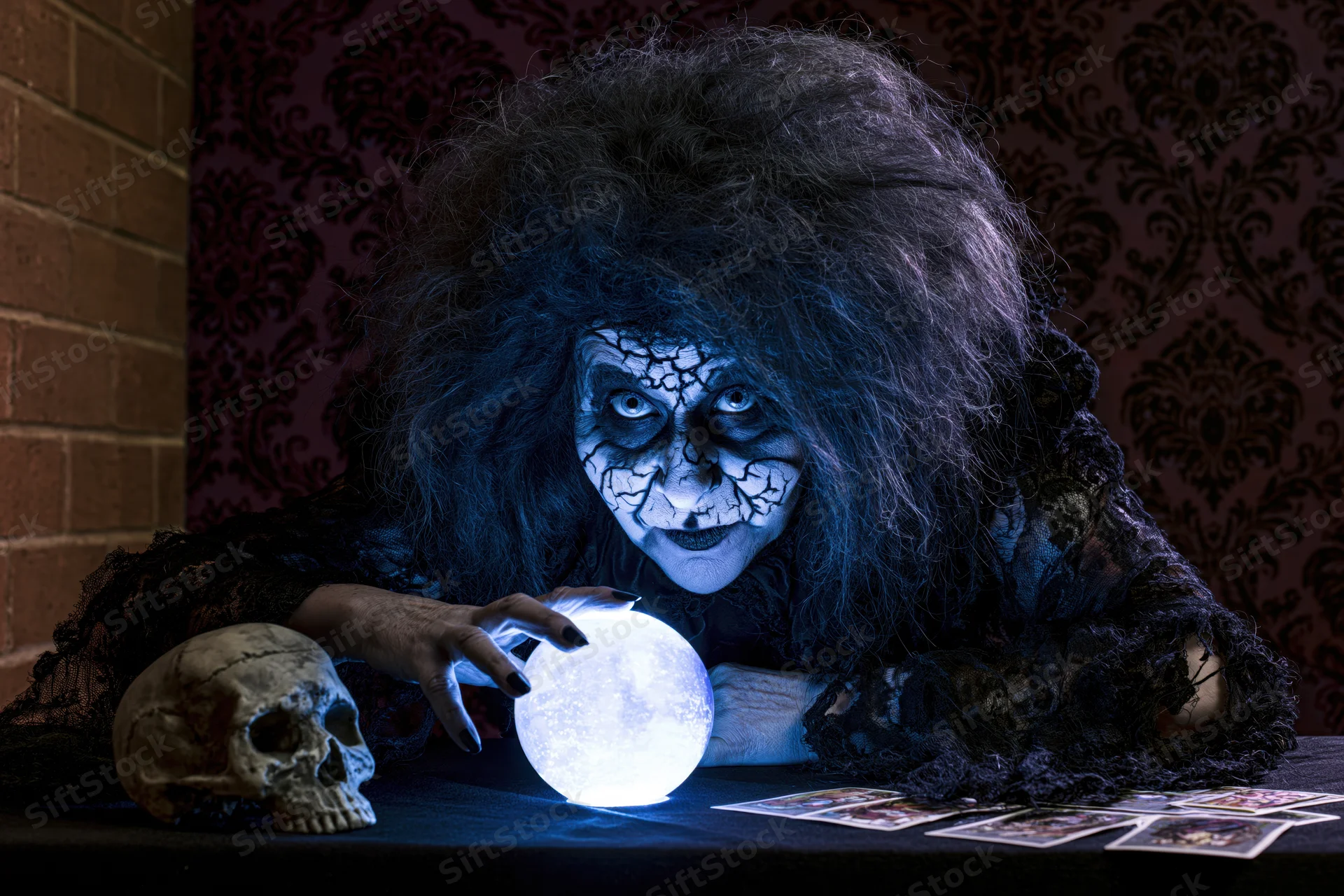 Witch fortune teller with cracked white makeup leaning over glowing crystal ball and tarot cards on dark table