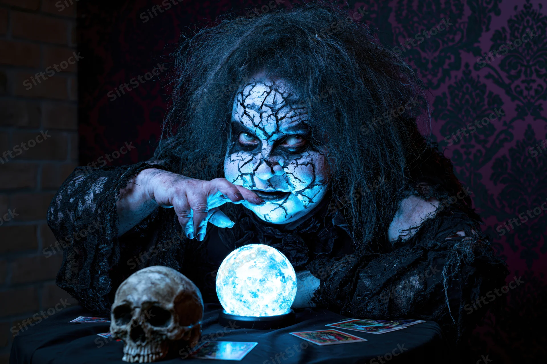 Witch fortune teller with cracked white face makeup leaning over glowing crystal ball and skull on dark table