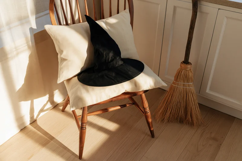 Witch hat resting on a cream linen pillow atop a wooden spindle-back chair beside a broom in a sunlit kitchen