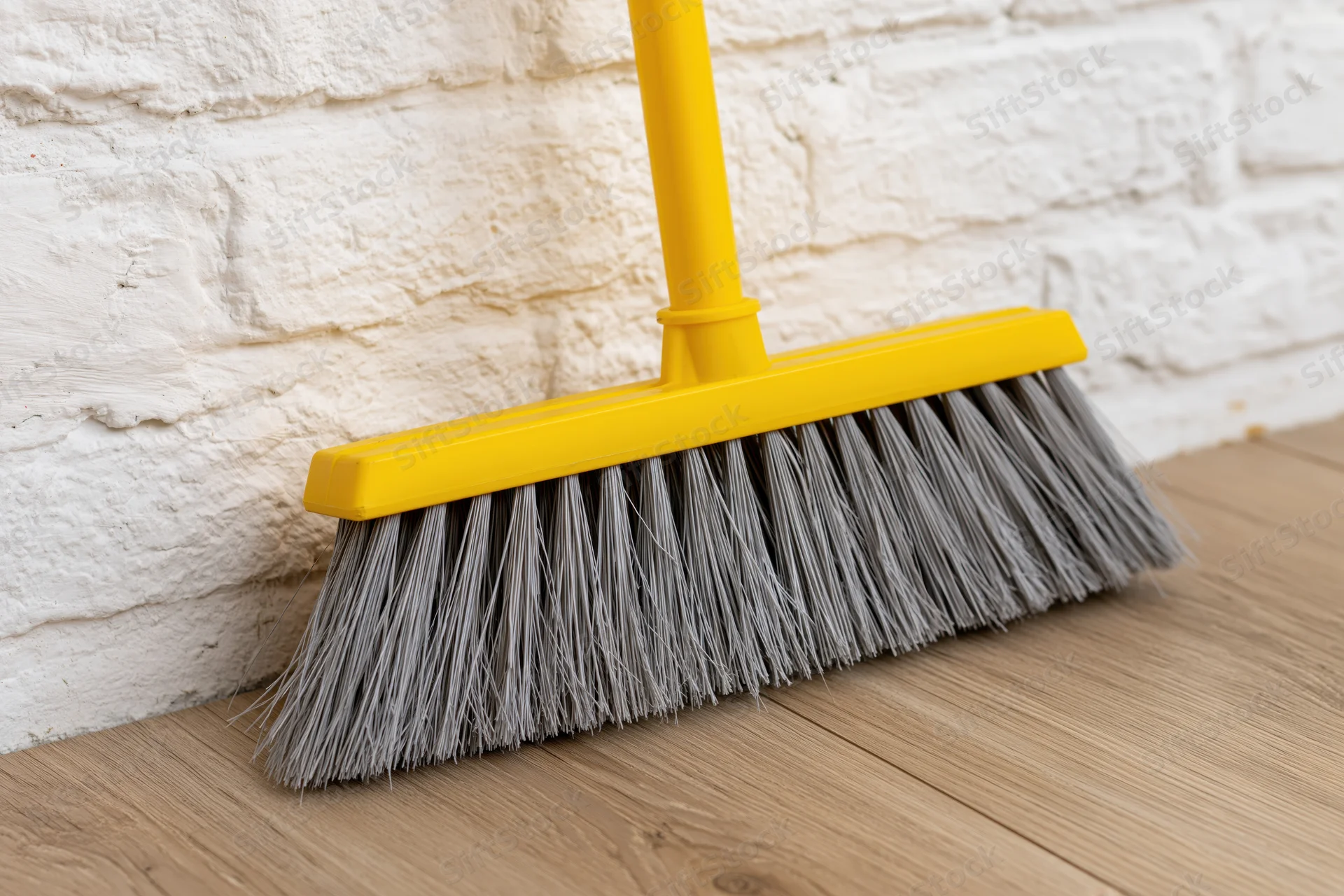 Yellow plastic broom with grey synthetic bristles leaning against white brick wall on wooden floor
