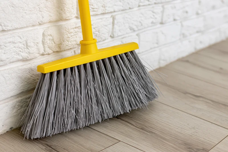 Yellow plastic broom with grey synthetic bristles leaning against white brick wall on wooden floor