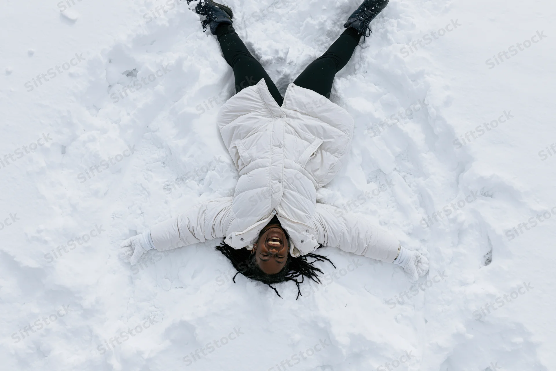 Young black woman lying in deep snow making a snow angel, smiling, overhead view