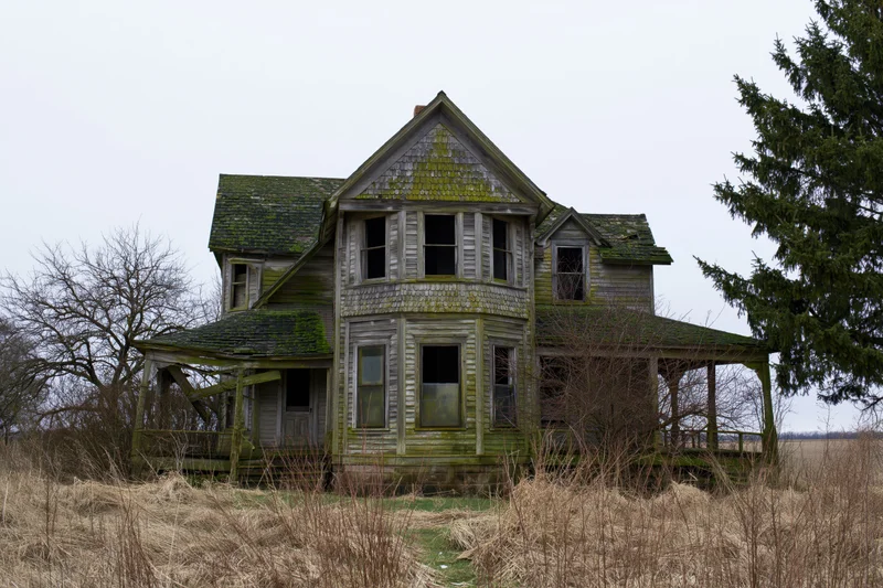 Abandoned two-story victorian farmhouse with collapsing front porch surrounded by overgrown field and bare trees