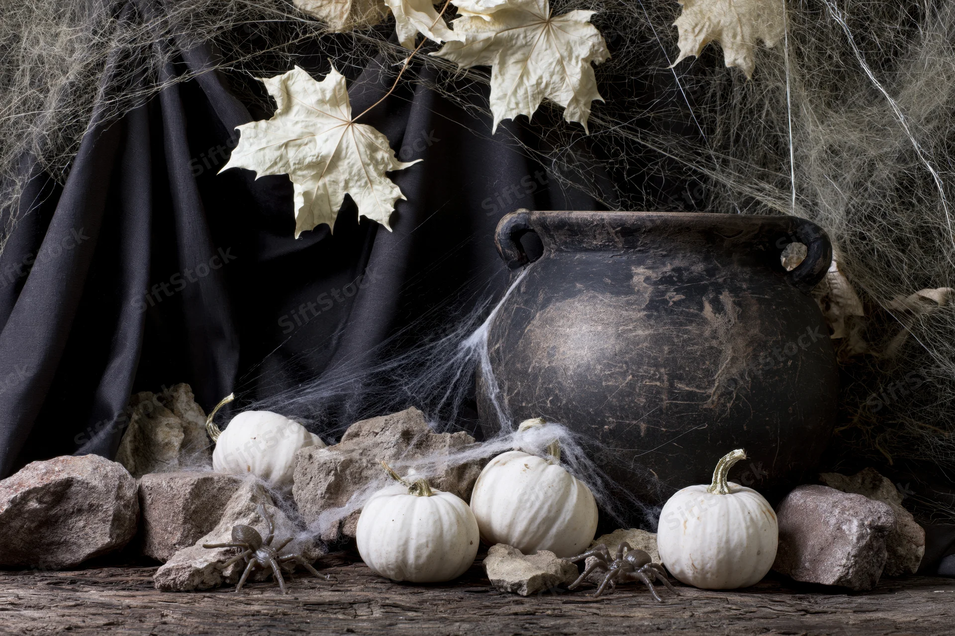Aged dark clay cauldron surrounded by white pumpkins, rocks, and cobwebs on a rough wooden surface