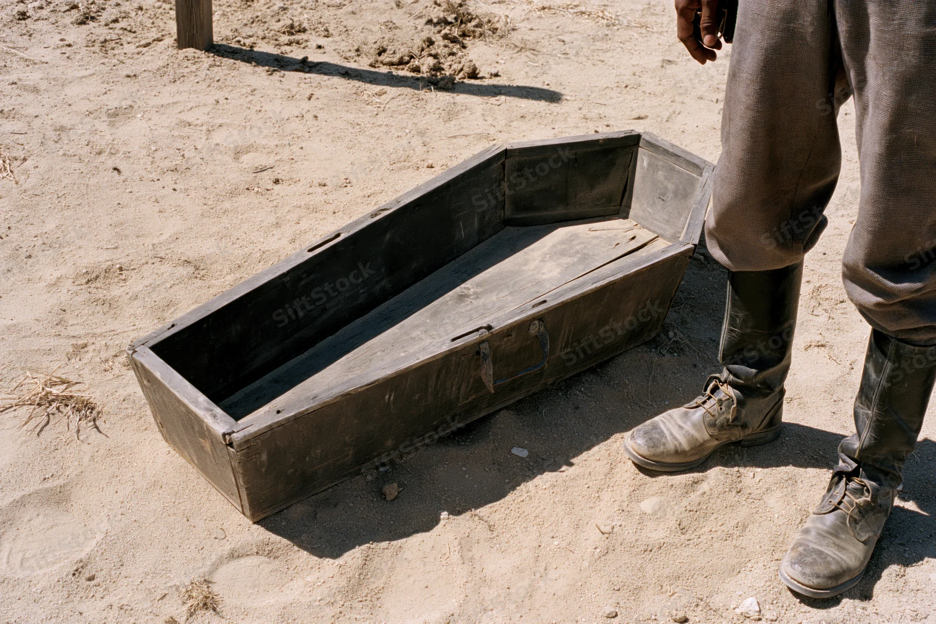 Open weathered wooden coffin on dry dusty ground beside a man in boots and trousers