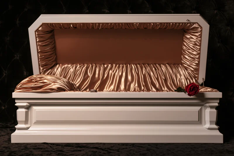 Open white wooden casket with bronze satin interior lining against dark tufted background