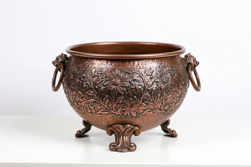 Antique hammered copper planter pot with floral embossing and lion handles on white surface