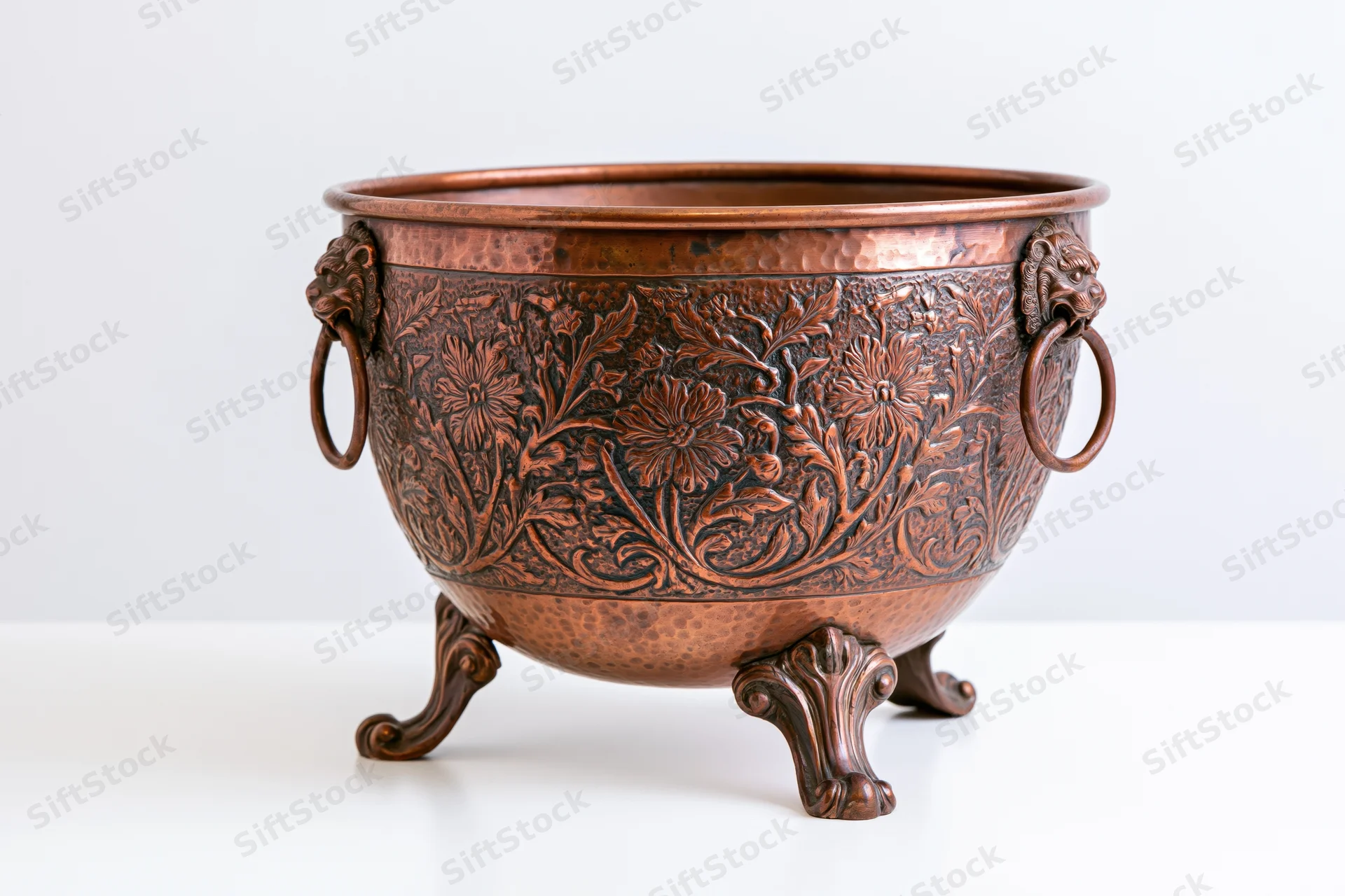 Antique hammered copper planter with floral embossing and lion ring handles on white surface