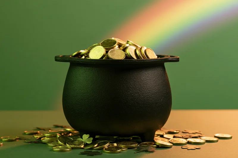 Black cauldron overflowing with gold coins and shamrocks on green surface with rainbow in background