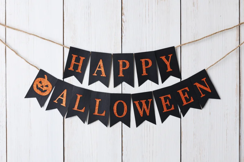 Black paper pennant banner spelling happy halloween with orange glitter text and pumpkin symbol hanging on twine against white wooden wall