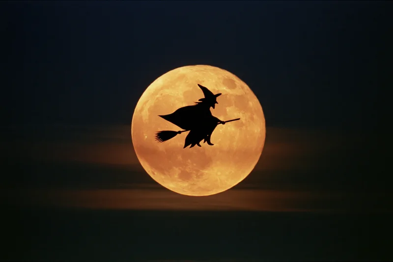 Witch flying on broomstick silhouetted against full orange moon in dark sky