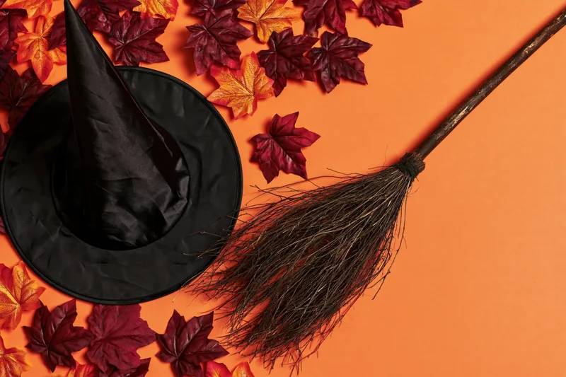 Black witch hat and rustic broom arranged with autumn leaves on orange background
