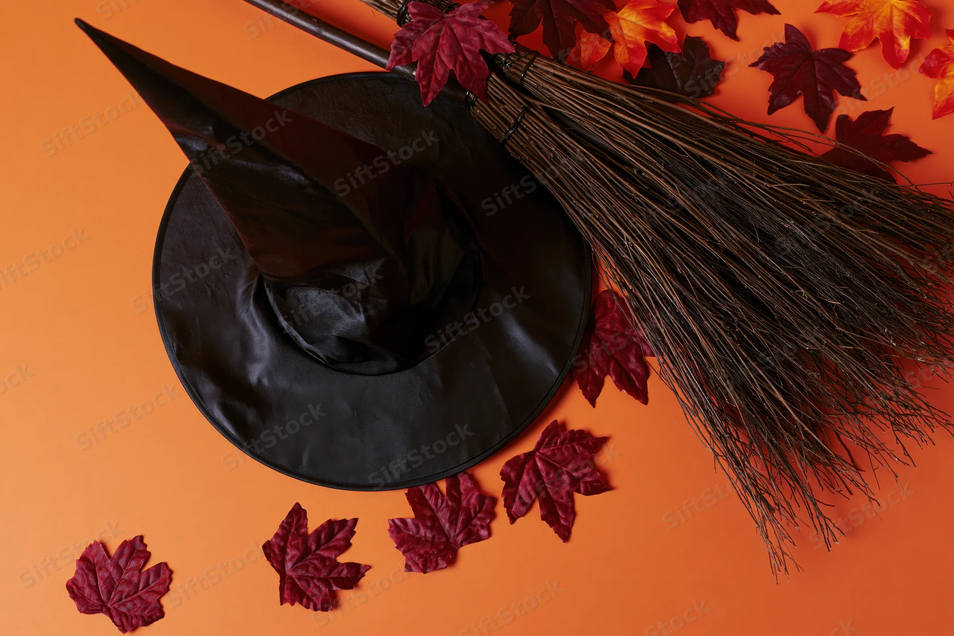 Black witch hat and rustic broom arranged with red maple leaves on orange background
