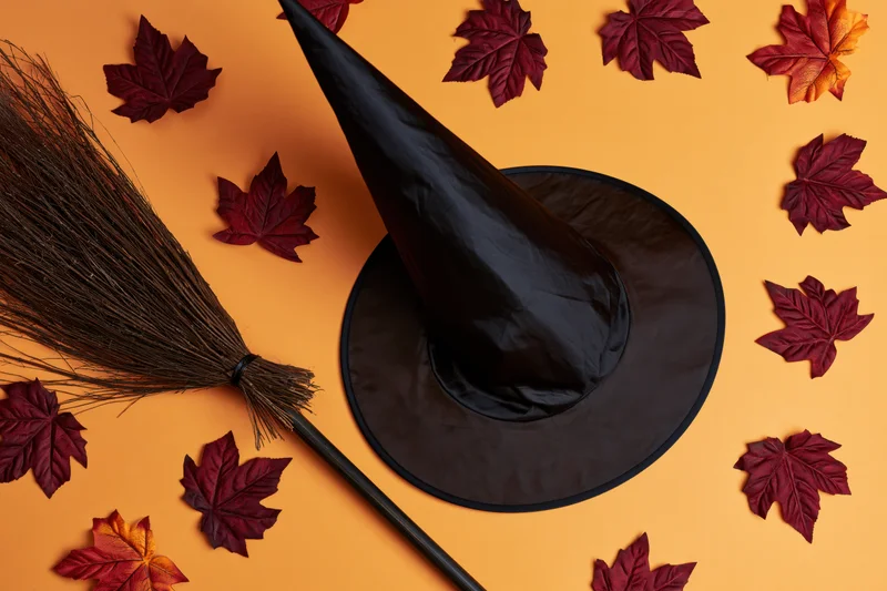 Black witch hat and rustic broom arranged with autumn leaves on orange background