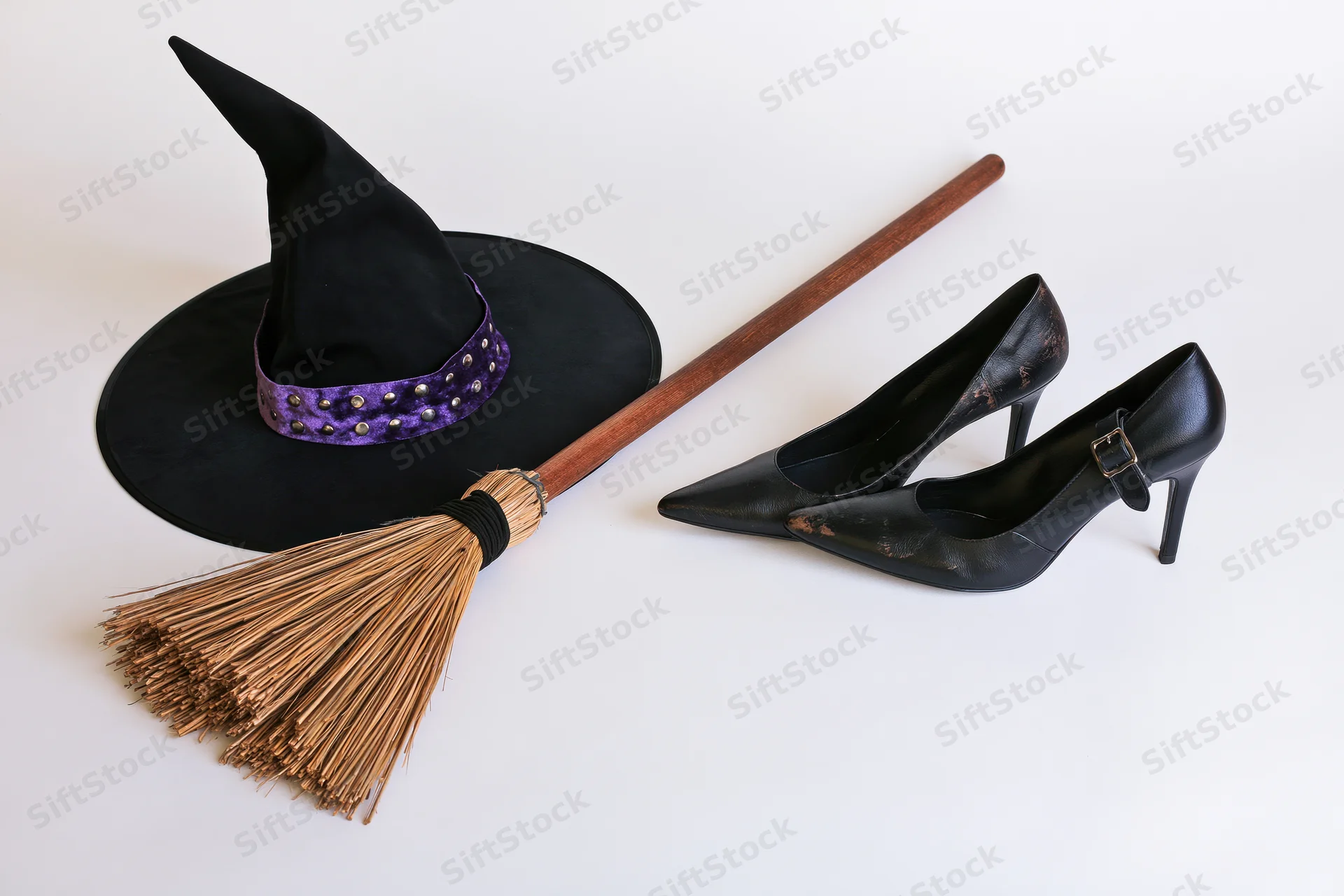 Witch hat with purple velvet band and metal studs, wooden broom, and black high heels arranged on white surface