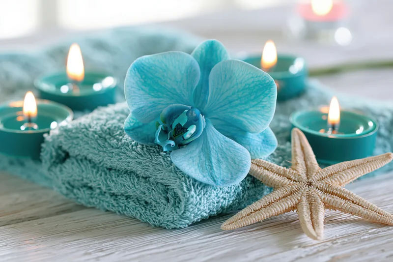 Blue orchid resting on folded aqua terry towel with lit teal candles and starfish on wooden surface