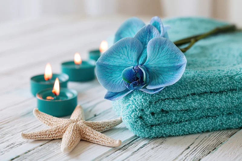 Blue orchid resting on folded aqua terry towel beside lit teal candles and starfish on wooden surface