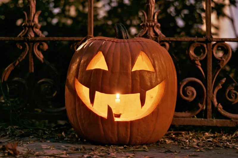 Carved pumpkin glowing with candlelight on a stone path beside a wrought iron fence at night