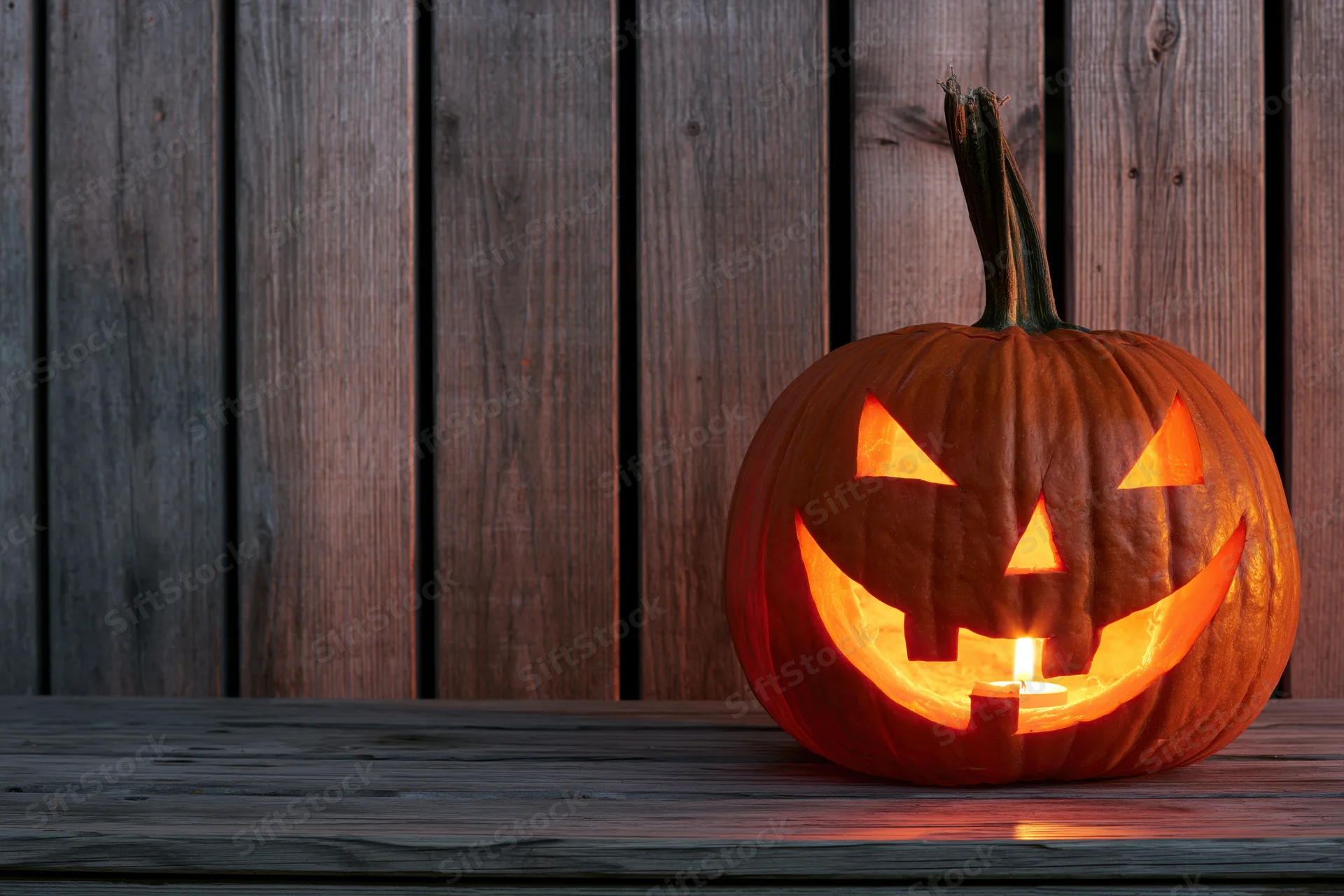 Carved pumpkin glowing with candlelight on wooden table against fence