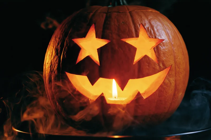 Carved pumpkin with star-shaped eyes and jagged smile glowing from within candlelight, surrounded by smoke on dark background
