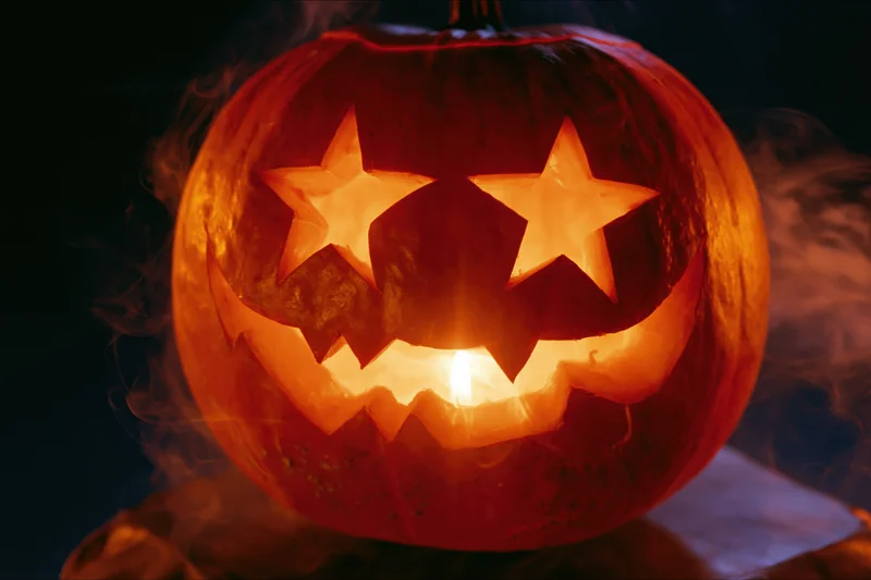 Carved pumpkin with star-shaped eyes and jagged grin glowing from within at night, surrounded by smoke
