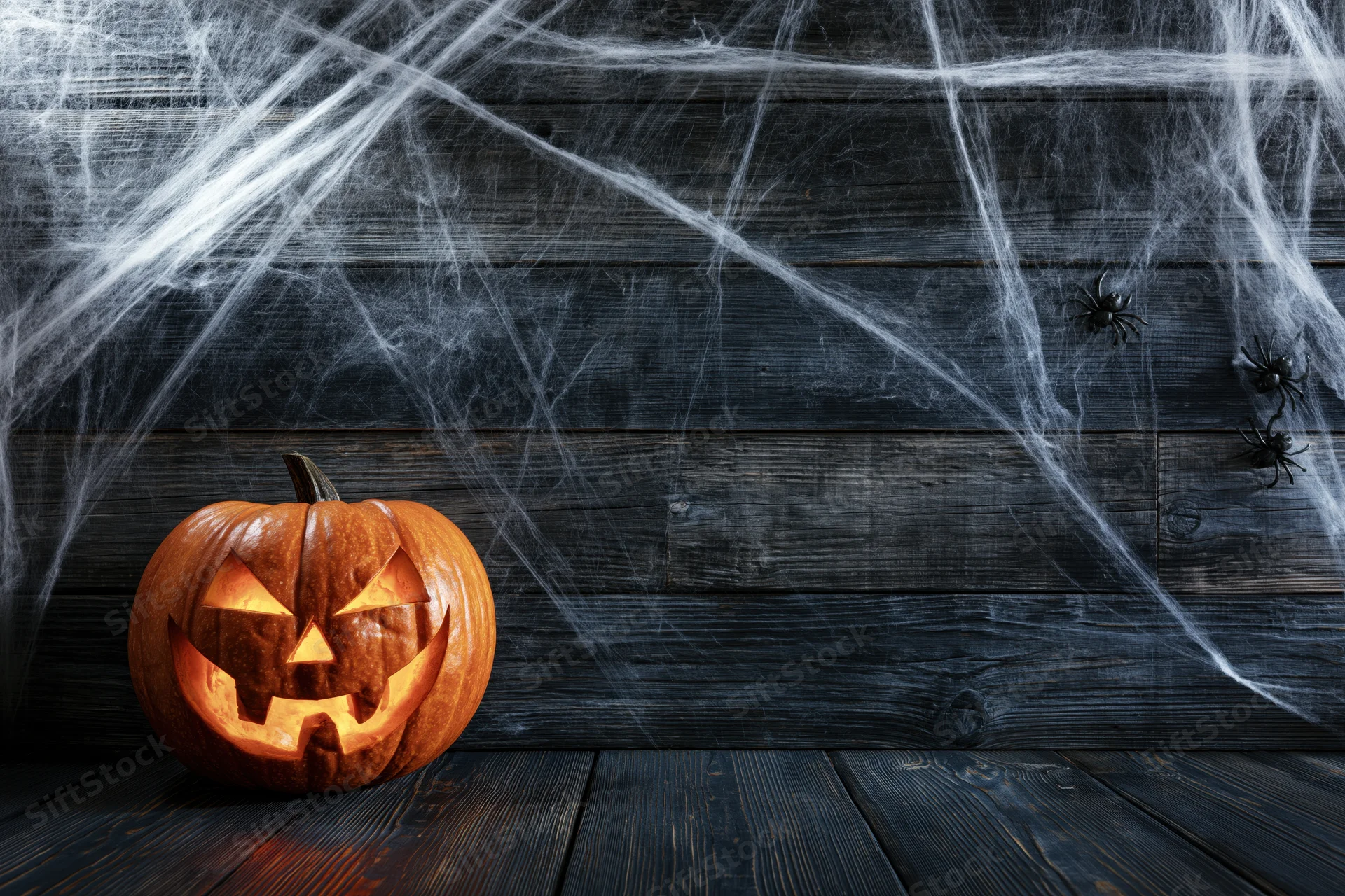 Carved jack-o-lantern glowing on dark wooden floor with spiderwebs and spiders