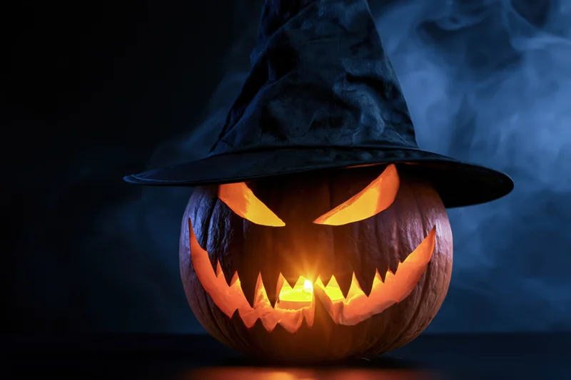 Carved jack-o-lantern with glowing eyes and jagged grin wearing a black witch hat against a smoky dark background