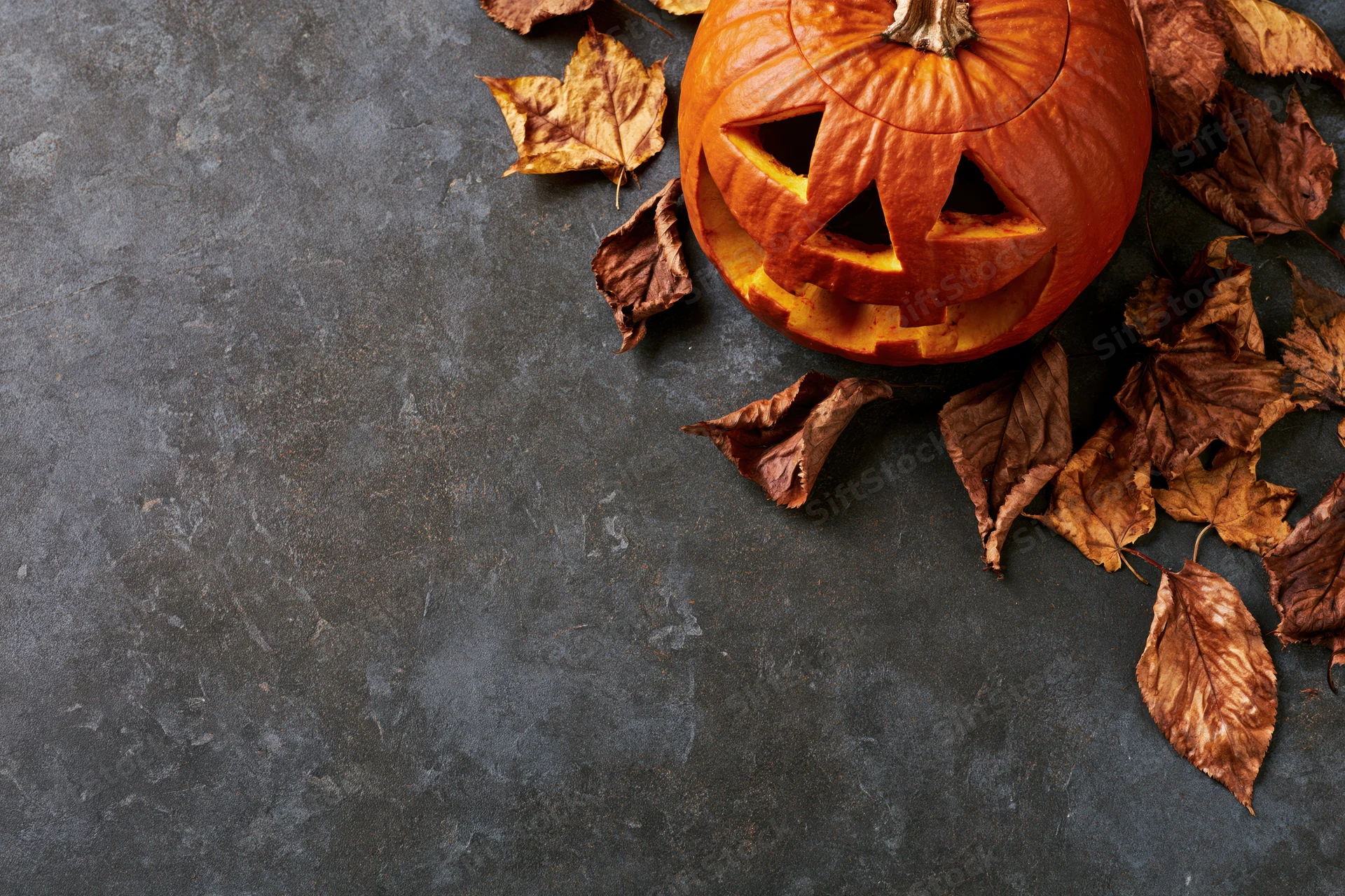 Carved pumpkin with triangular eyes and dry autumn leaves on dark stone surface