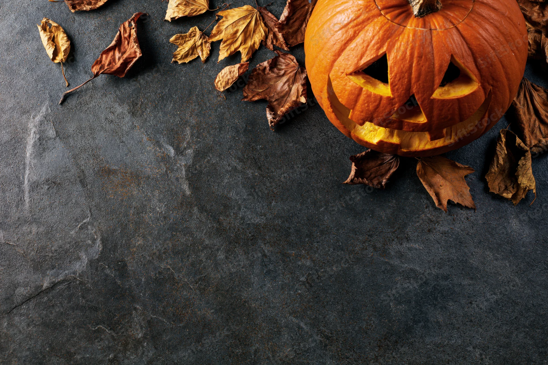 Carved pumpkin with triangular eyes and dried leaves on dark stone surface