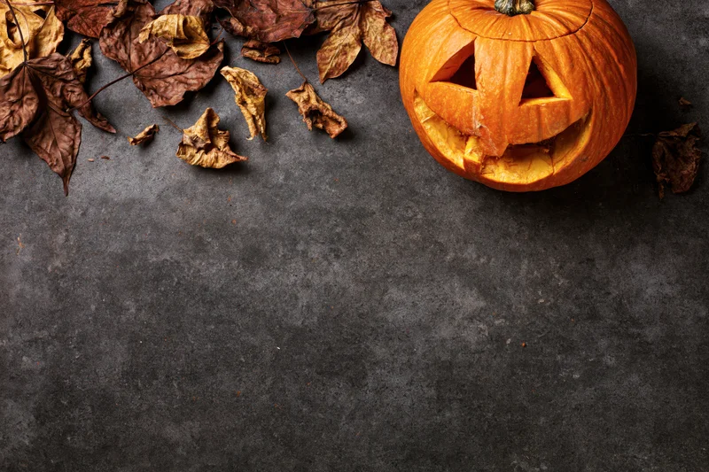 Carved pumpkin with triangular eyes and dried leaves arranged on dark concrete surface