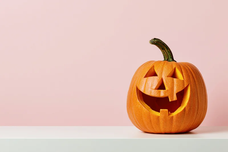 Carved pumpkin with grinning face on a white surface against pink background