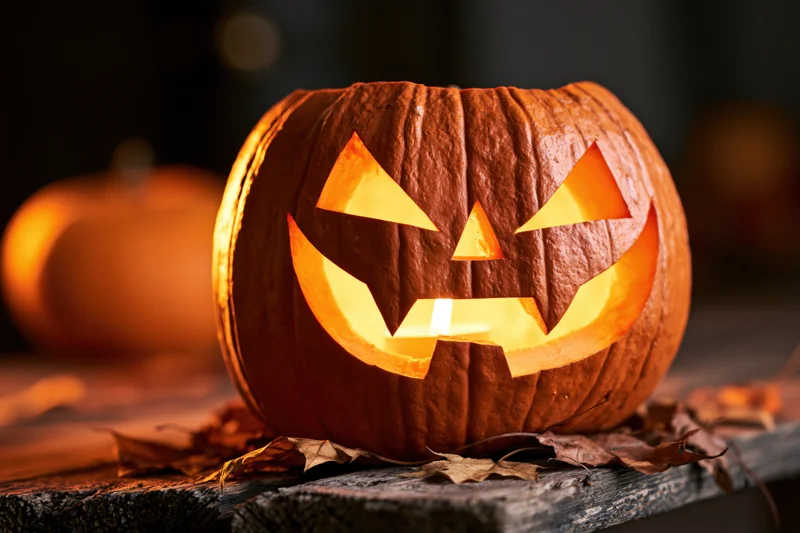Carved pumpkin with jagged grinning face glowing on wooden table surrounded by dry leaves