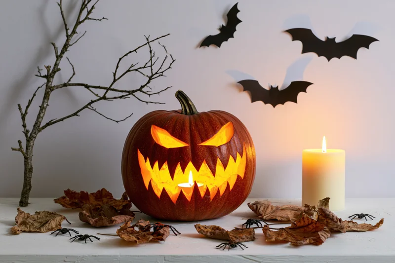 Carved jack-o-lantern with jagged grin lit by candle on white table surrounded by bats, leaves, and spiders