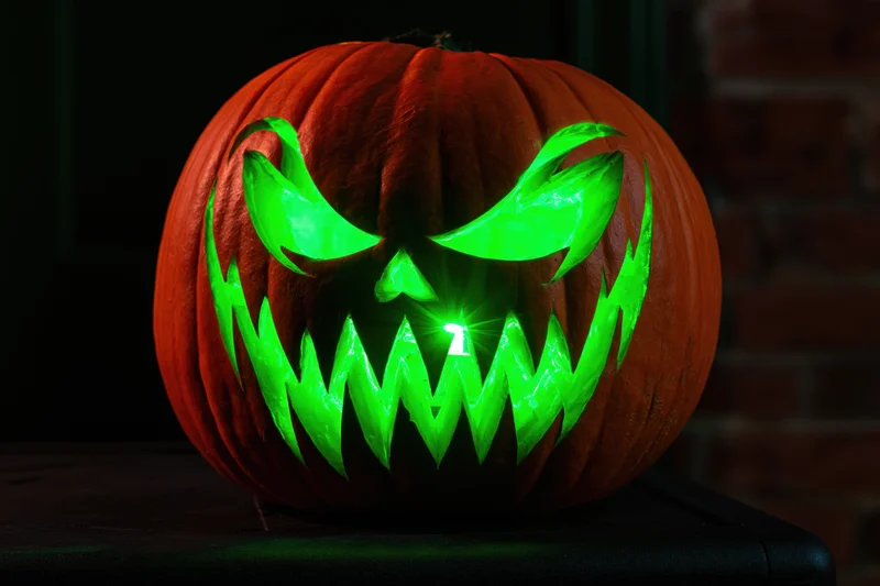 Carved pumpkin with glowing green evil face on dark table near brick wall