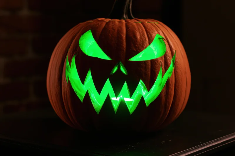 Carved pumpkin with angular glowing green face on dark table near brick wall