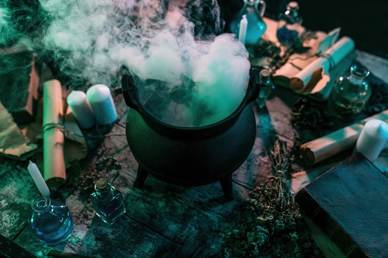 Black cast iron cauldron billowing thick white smoke on a wooden table surrounded by scrolls, herbs, candles and glass bottles