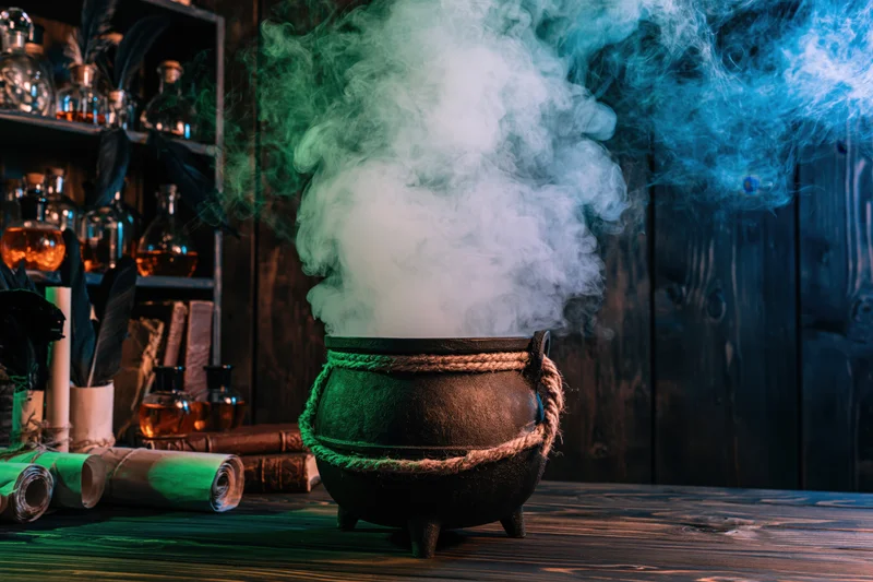 Cast iron cauldron emitting green and blue smoke on wooden table surrounded by potion bottles and scrolls